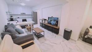 Un salon moderne avec un canapé, une table basse et un téléviseur. On aperçoit, au fond, une cuisine avec des meubles blancs et des appareils électroménagers.