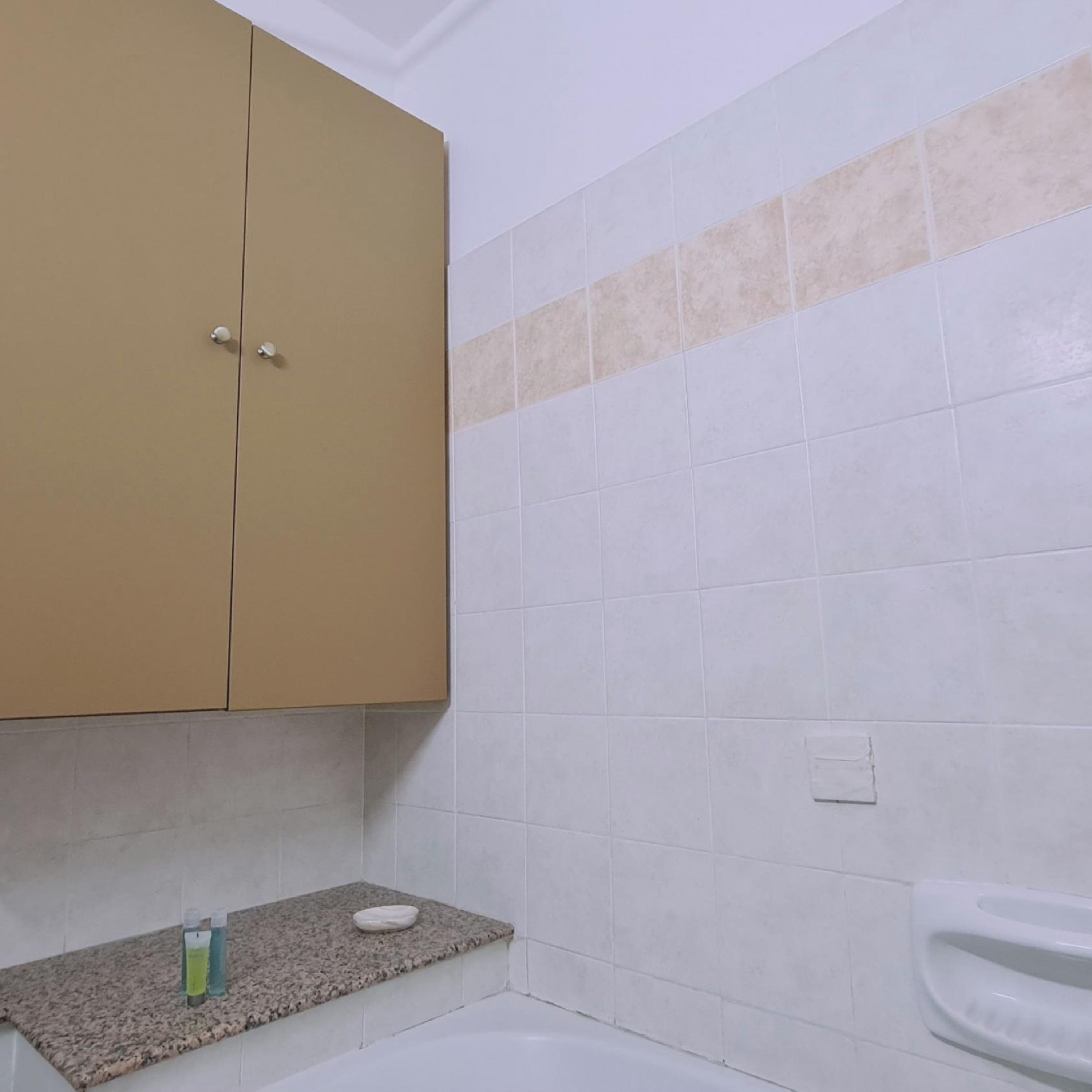 Un baño con un mueble color beige, una encimera de granito, un inodoro blanco y una pared con azulejos color beige.