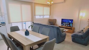 Un salon moderne avec une table à manger en bois, des chaises grises, un canapé bleu, un téléviseur posé sur un meuble en bois et une plante.