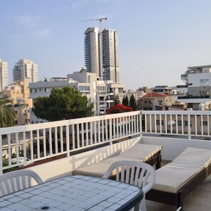Une terrasse sur le toit, avec une table et des chaises, une rambarde blanche, et une vue sur les bâtiments de la ville et une grue de construction en arrière-plan.