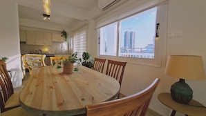 A dining area with a wooden table, chairs, and a lamp. There is a window with a view of buildings and a plant on the table. - 83 Lim2 - Stay Naturaleza Penthouse (Limassol)
