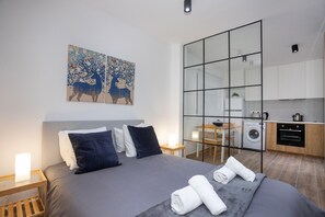 A modern hotel room with a large bed, a small table, a wall-mounted TV, and a kitchen area visible through a glass partition. - 89 LIM - Stay Zen Studio (Limassol)