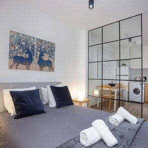 A modern bedroom with a large bed, two wall-mounted paintings, a small bedside table, and a view into a kitchen area. - 89 LIM - Stay Zen Studio (Limassol)