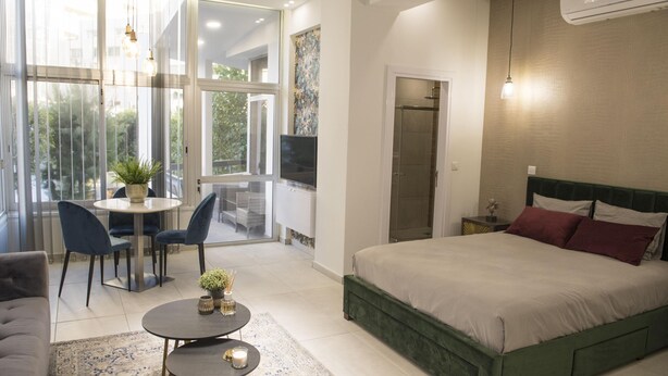 Une chambre moderne avec un grand lit, un coin repas avec des chaises, une petite table agrémentée d’une plante et une grande fenêtre donnant sur l’extérieur.