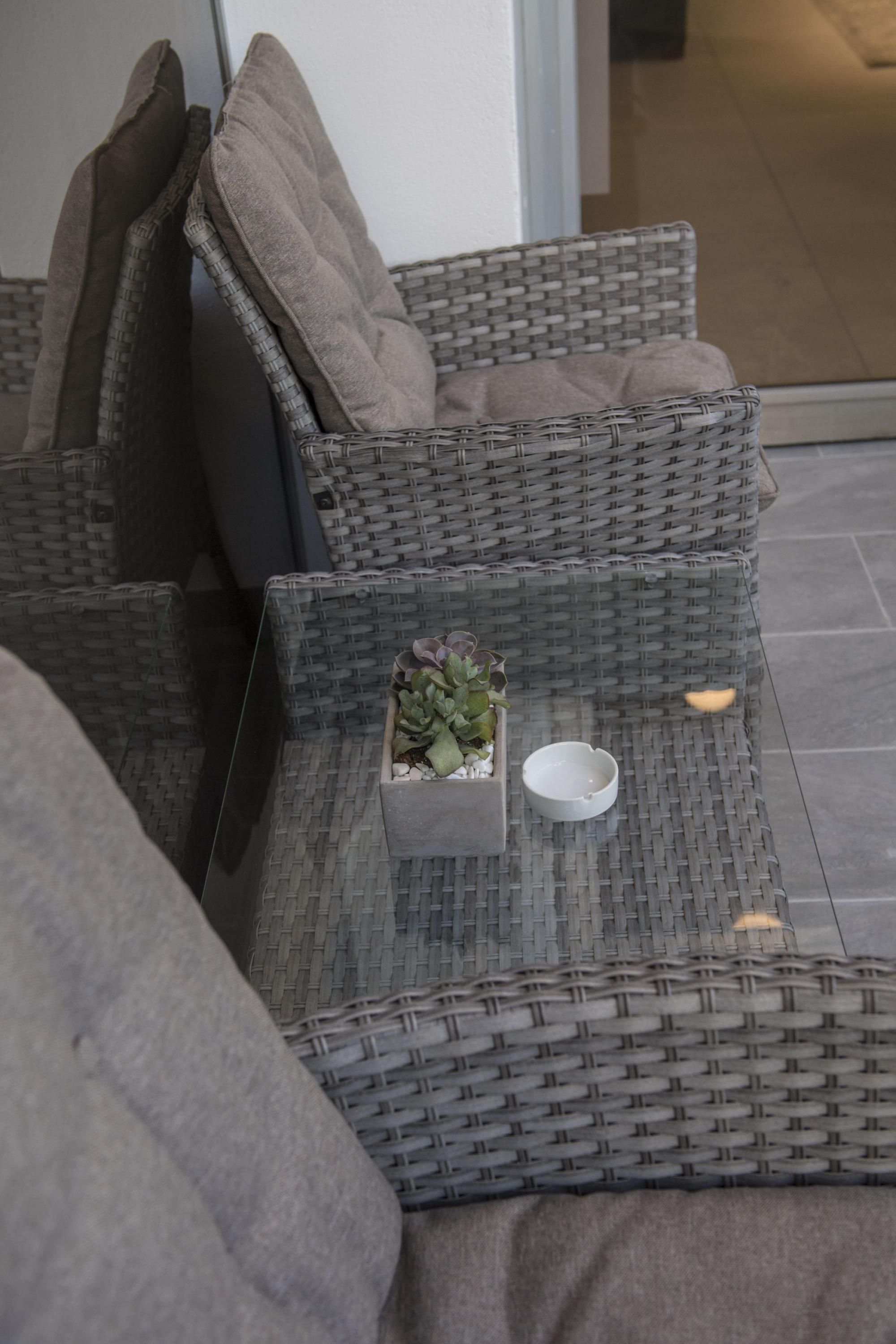 A wicker sofa with cushions, a small potted plant, and a white ashtray on a woven floor.