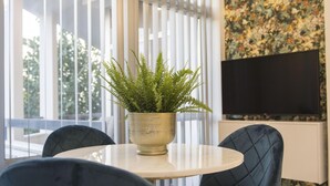 Un salon moderne avec une table ronde, deux fauteuils bleus, une fougère en pot, un téléviseur à écran plat et un mur orné d’un motif floral.