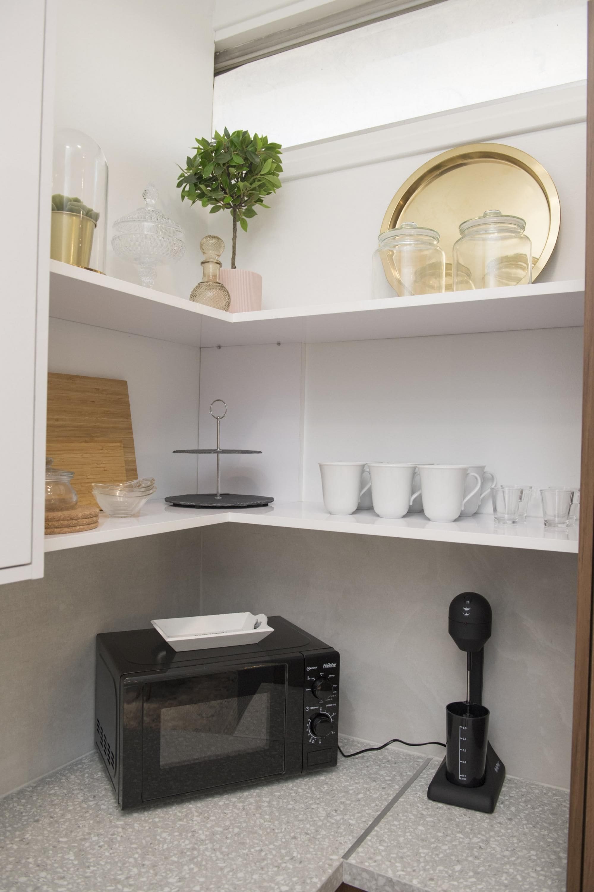 A compact kitchen with a microwave, coffee maker, and shelves with jars and cups.