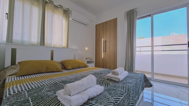 Un dormitorio moderno con una cama grande, dos almohadas, una mesita de noche con lámpara, un armario de madera y vistas al exterior.