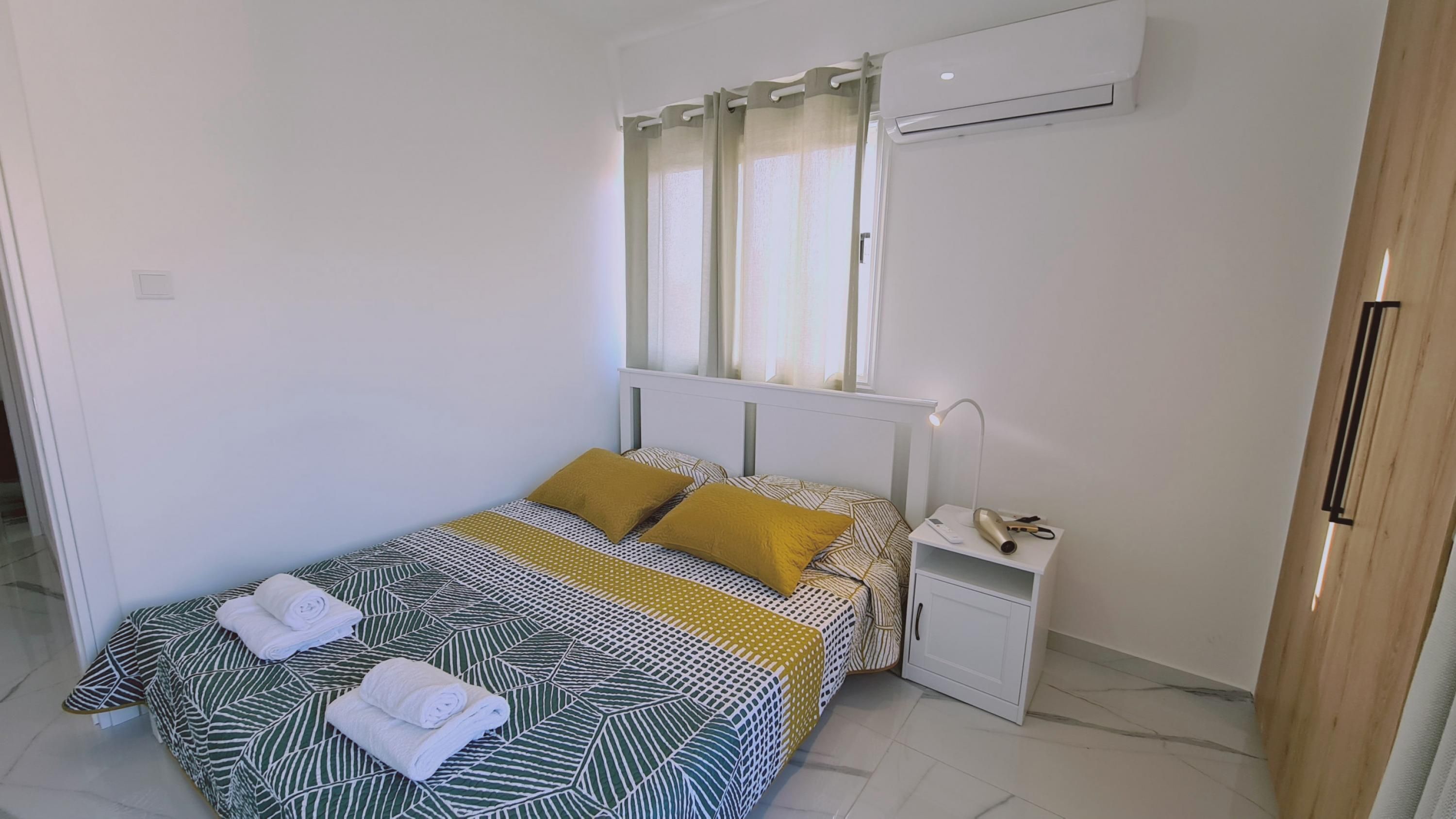 A neatly made bed with a patterned bedspread and two pillows, a bedside table with a lamp and a hairdryer, and an air conditioning unit on the wall.