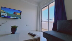 Una sala de estar moderna con un televisor de pantalla plana montado en la pared mostrando la interfaz de Netflix, un control remoto sobre una mesa blanca y una planta en maceta.