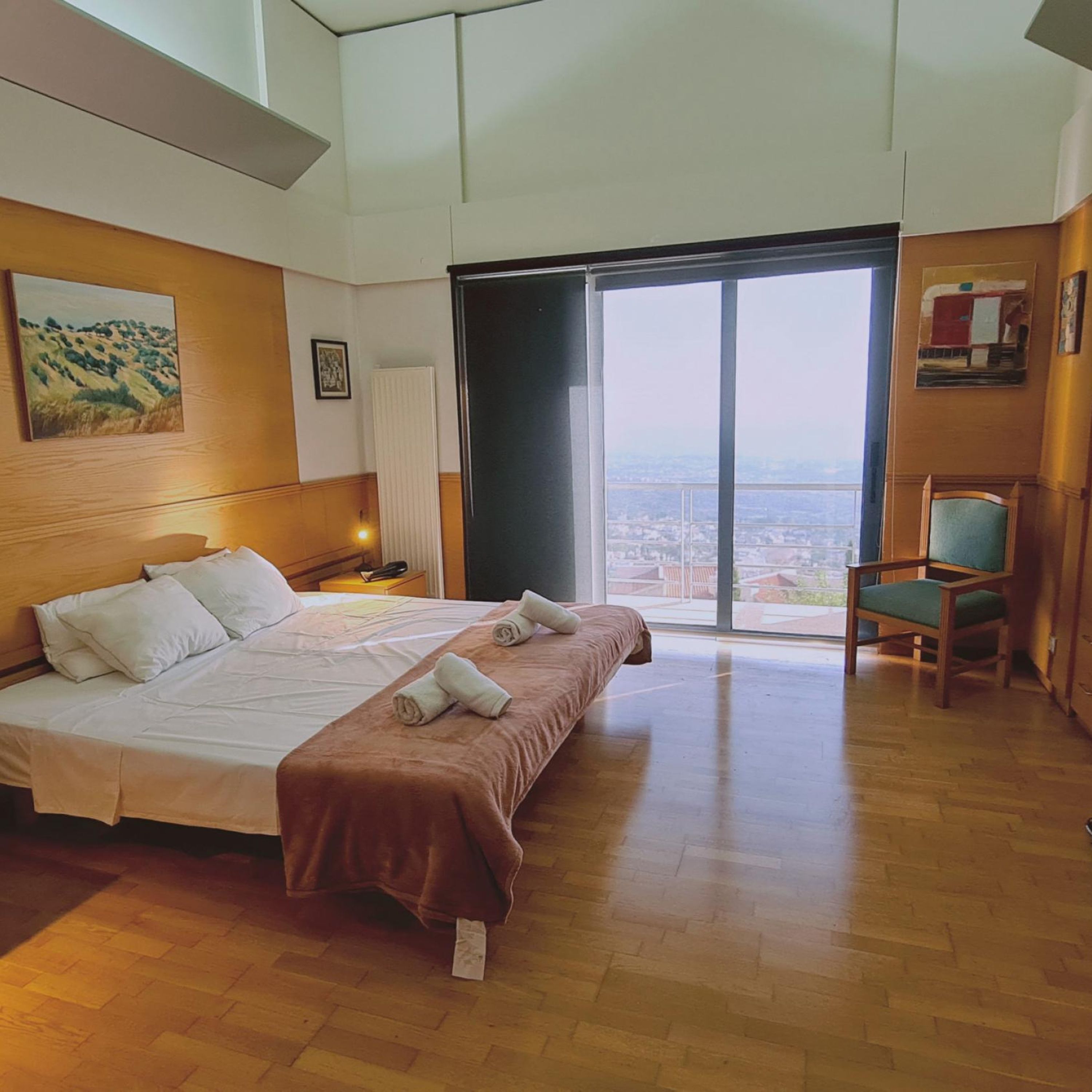 A hotel room with a large bed, a view through a sliding door, a chair, and a painting on the wall.