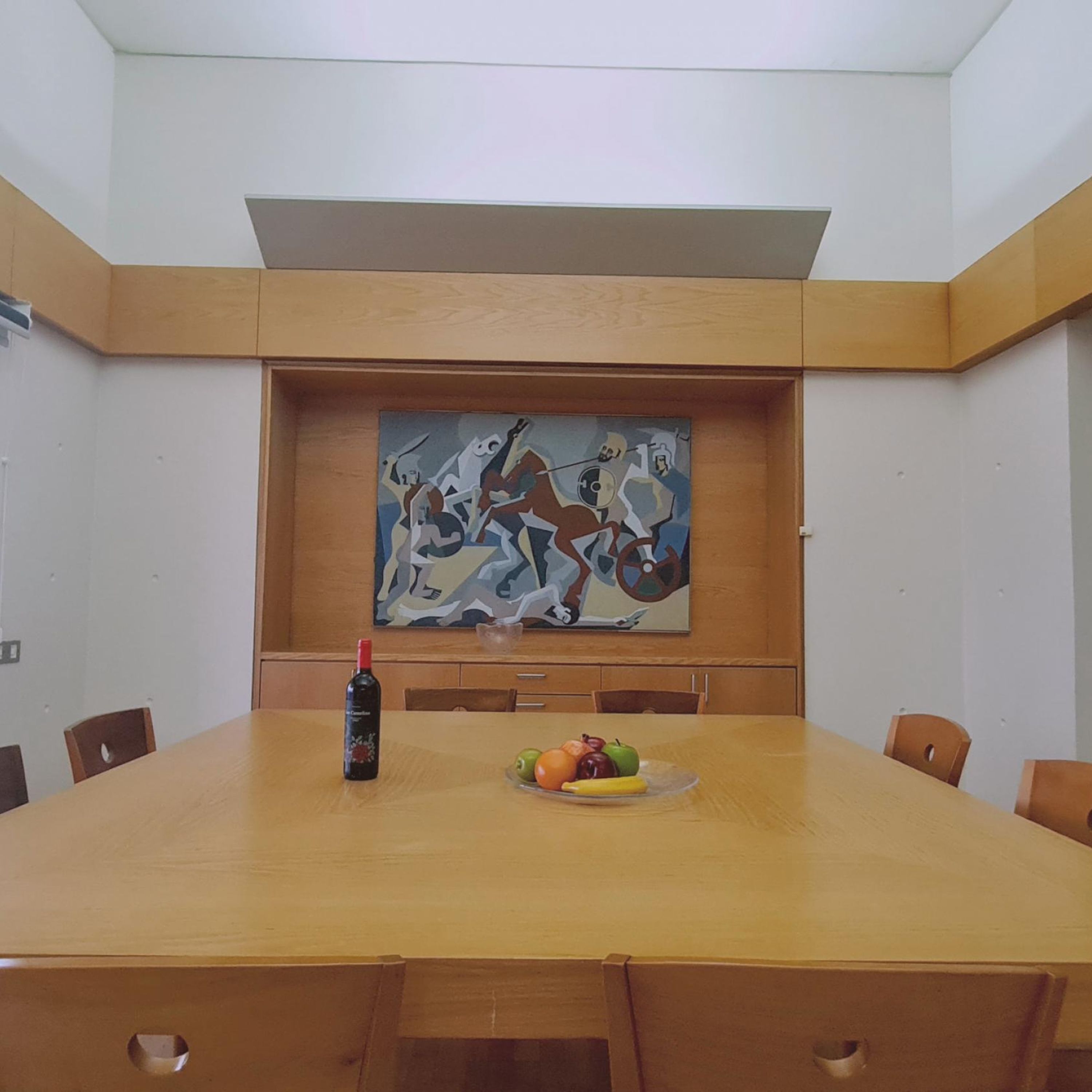 A conference room with a large table, chairs, a painting on the wall, and a bottle of wine on the table.
