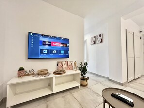Een moderne woonkamer met een wandtelevisie waarop een beginscherm staat, een witte console met decoratieve objecten, en een afstandsbediening op een dienblad.