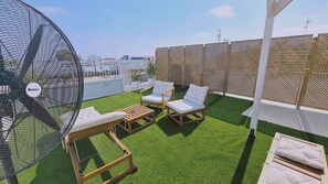 A rooftop terrace with artificial grass, two white cushioned chairs, a wooden table, and a large fan. - 84 AN 1 - Stay Napa Aura Apartment (Ayia Napa)