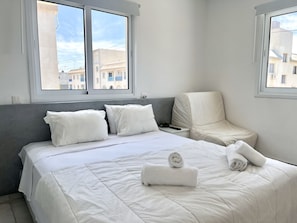A hotel room with a bed, a grey headboard, a white bedside table, and two windows. - 84 AN 1 - Stay Napa Aura Apartment (Ayia Napa)