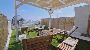Una terraza en la azotea con mobiliario de madera, un ventilador grande y vistas de edificios al fondo.