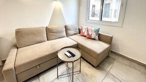 A living room with a beige sectional sofa, a small round table with a remote control, a wall-mounted light fixture, and a window with a view of buildings. - 84 AN 1 - Stay Napa Aura Apartment (Ayia Napa)