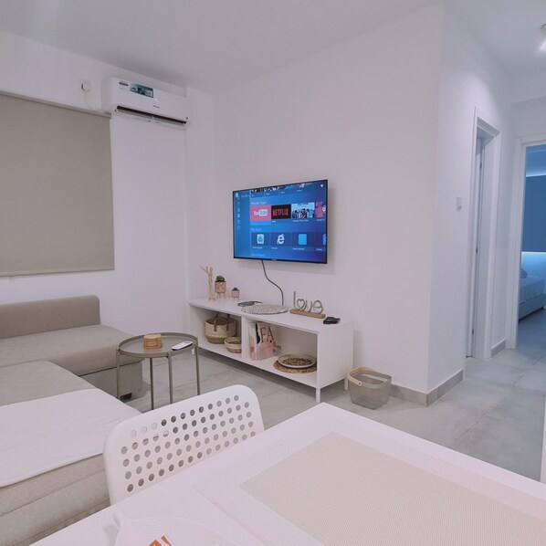 Una sala de estar moderna con un televisor de pantalla plana montado en la pared, un sofá, una mesita redonda y un aparador blanco con objetos decorativos.