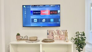 A flat-screen TV mounted on a wall displaying a media application interface with options like YouTube, Netflix, and MetaX TV. Below the TV, there's a white console with decorative items and a potted plant. - 84 AN 1 - Stay Napa Aura Apartment (Ayia Napa)