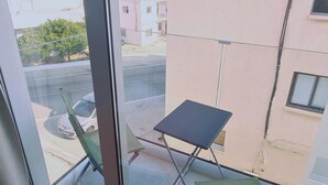A balcony with a small table and two chairs, overlooking a street with buildings and a car. - 100 AN - Napa Living 23 (Ayia Napa)