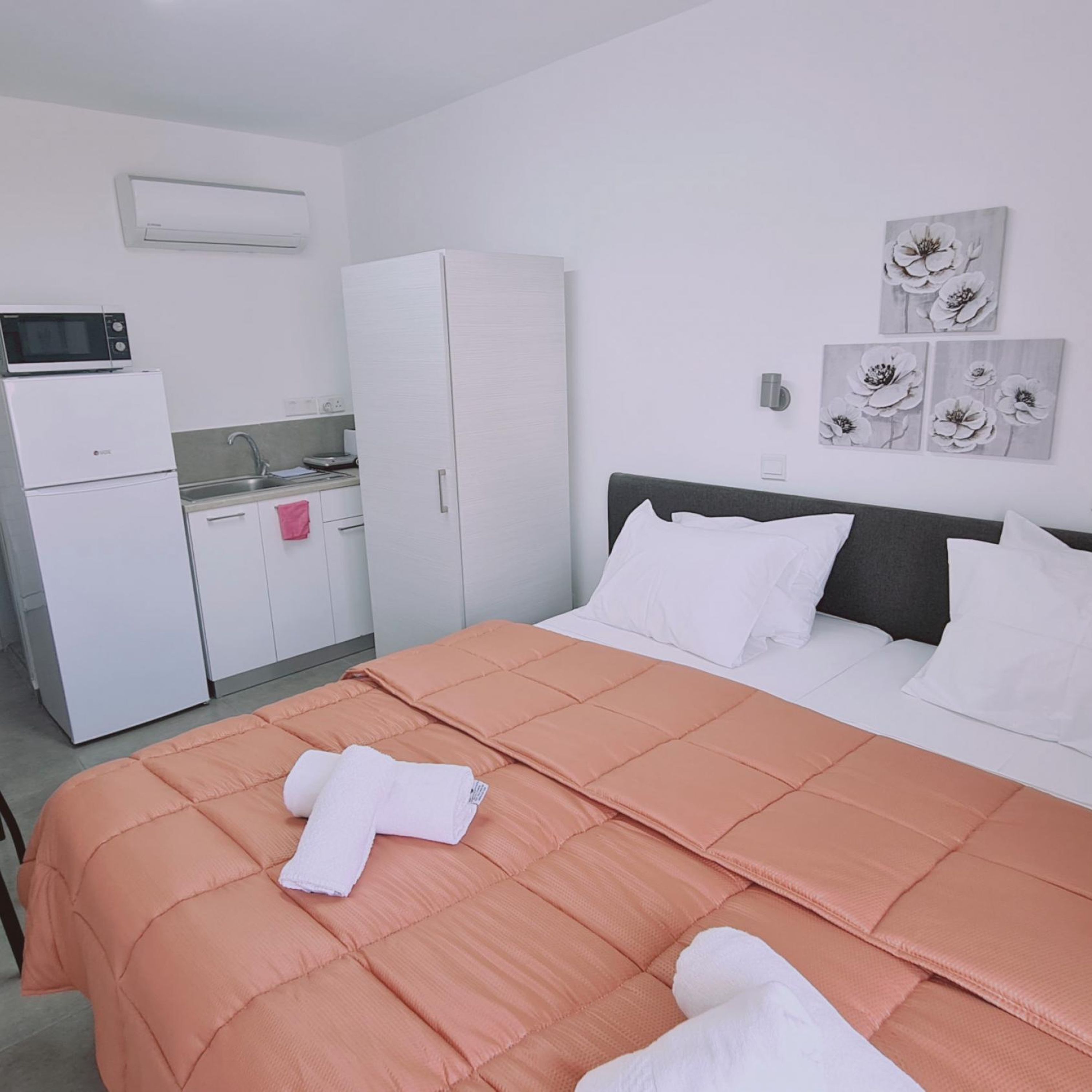 Ein Schlafzimmer mit einem Bett, weißen Kissen, einer pfirsichfarbenen Bettdecke, einem weißen Handtuch, einer Mikrowelle, einem Kühlschrank, einem Waschbecken und einem Schrank.