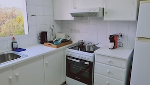 A compact kitchen with white cabinets, a stove, a sink, and a window with a view of trees. - 99 LIM - Stay Almeria Apartment (Limassol)
