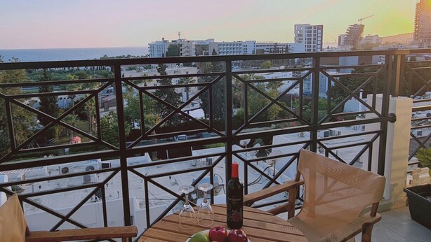 A balcony with a table set for two, featuring a bottle of wine, a plate with fruit, and a view of the cityscape and sea. - 99 LIM - Stay Almeria Apartment (Limassol)