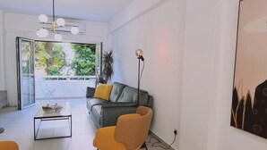 A modern living room with a grey sofa, yellow armchair, and a coffee table. There is a large window with a view of greenery outside. - 97 LIM - Stay Vital Elegance Apartment (Limassol)