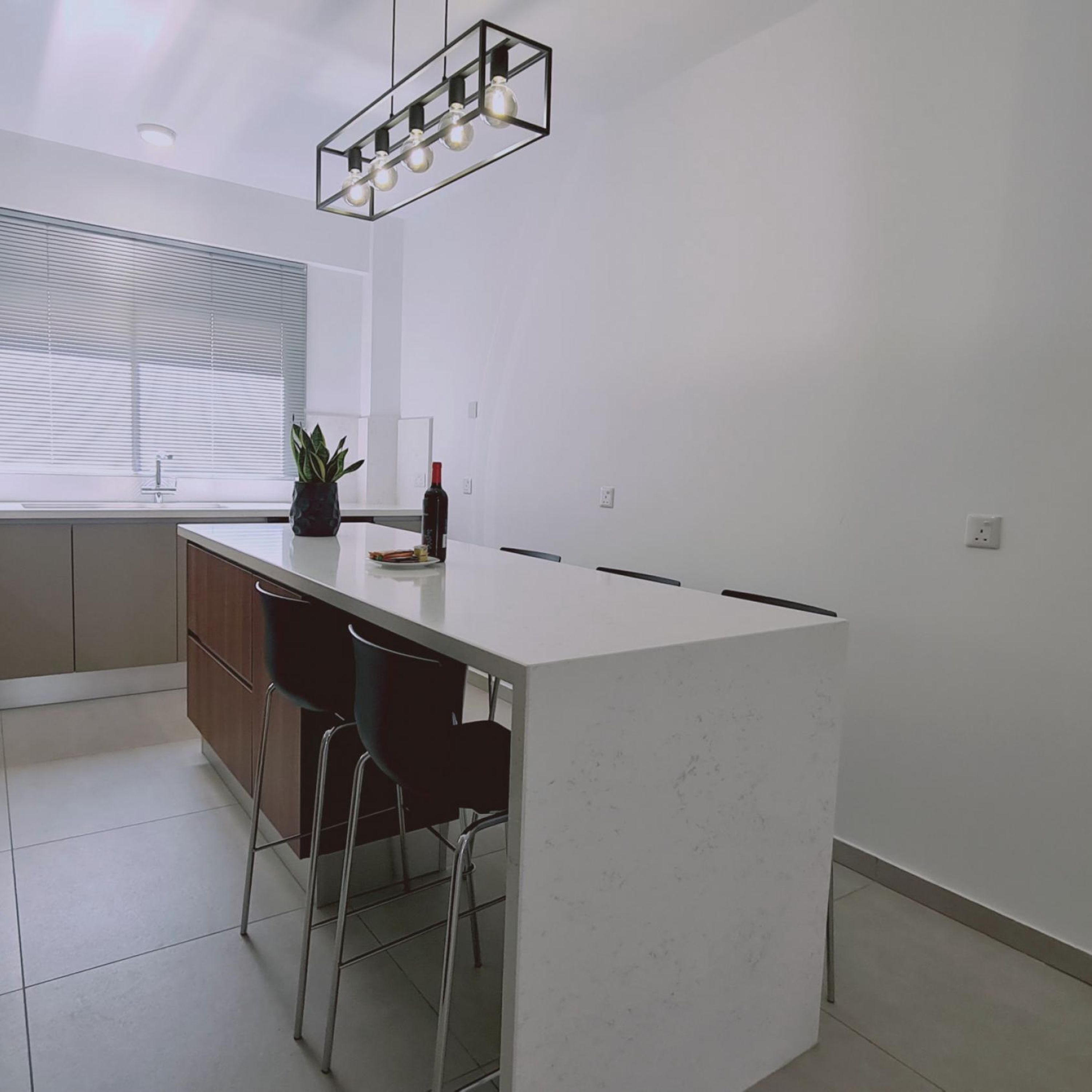 A modern kitchen with a white island, bar stools, a hanging light fixture, and a window with blinds.