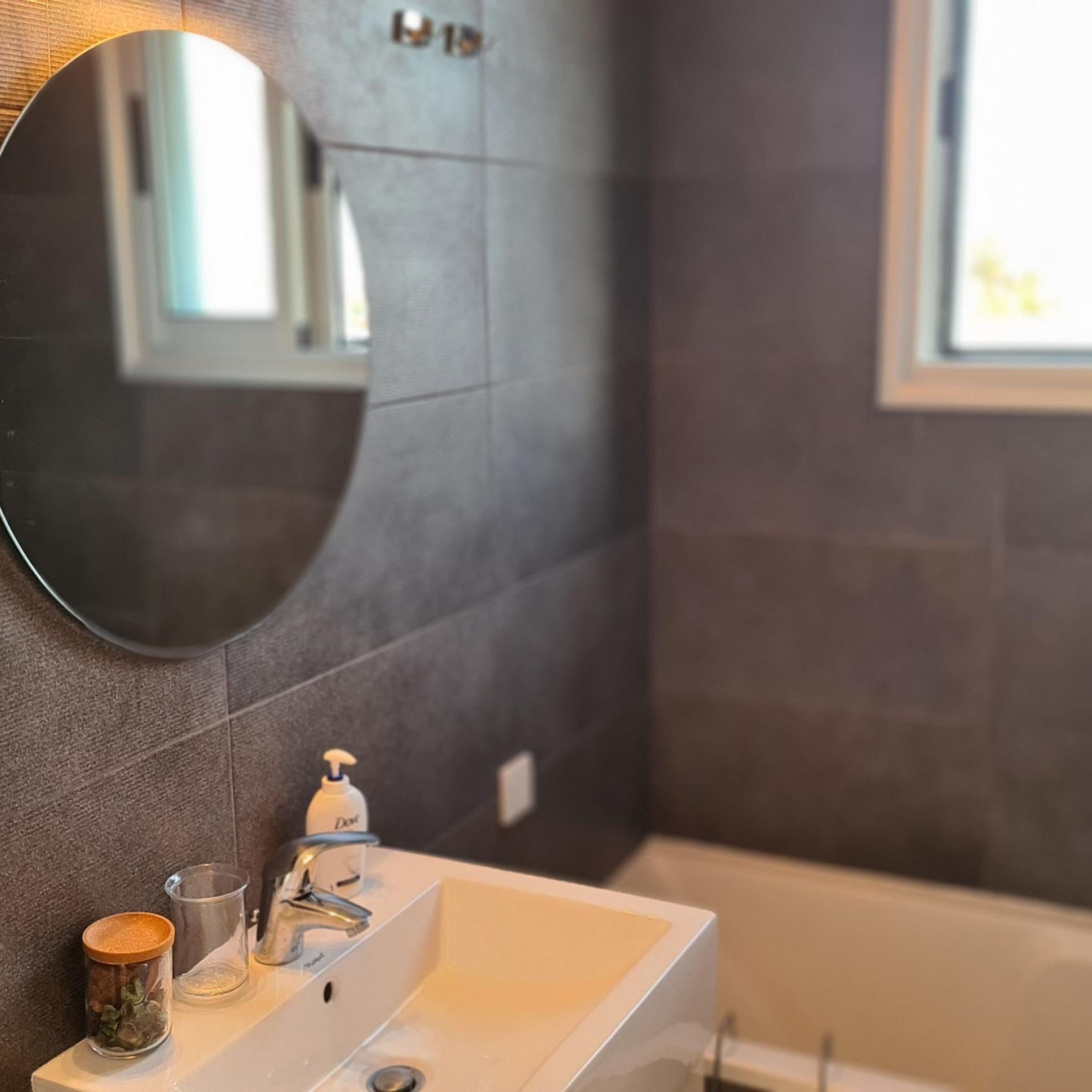 Een badkamer met een wit wastafelmeubel, een ronde spiegel en een raam.
