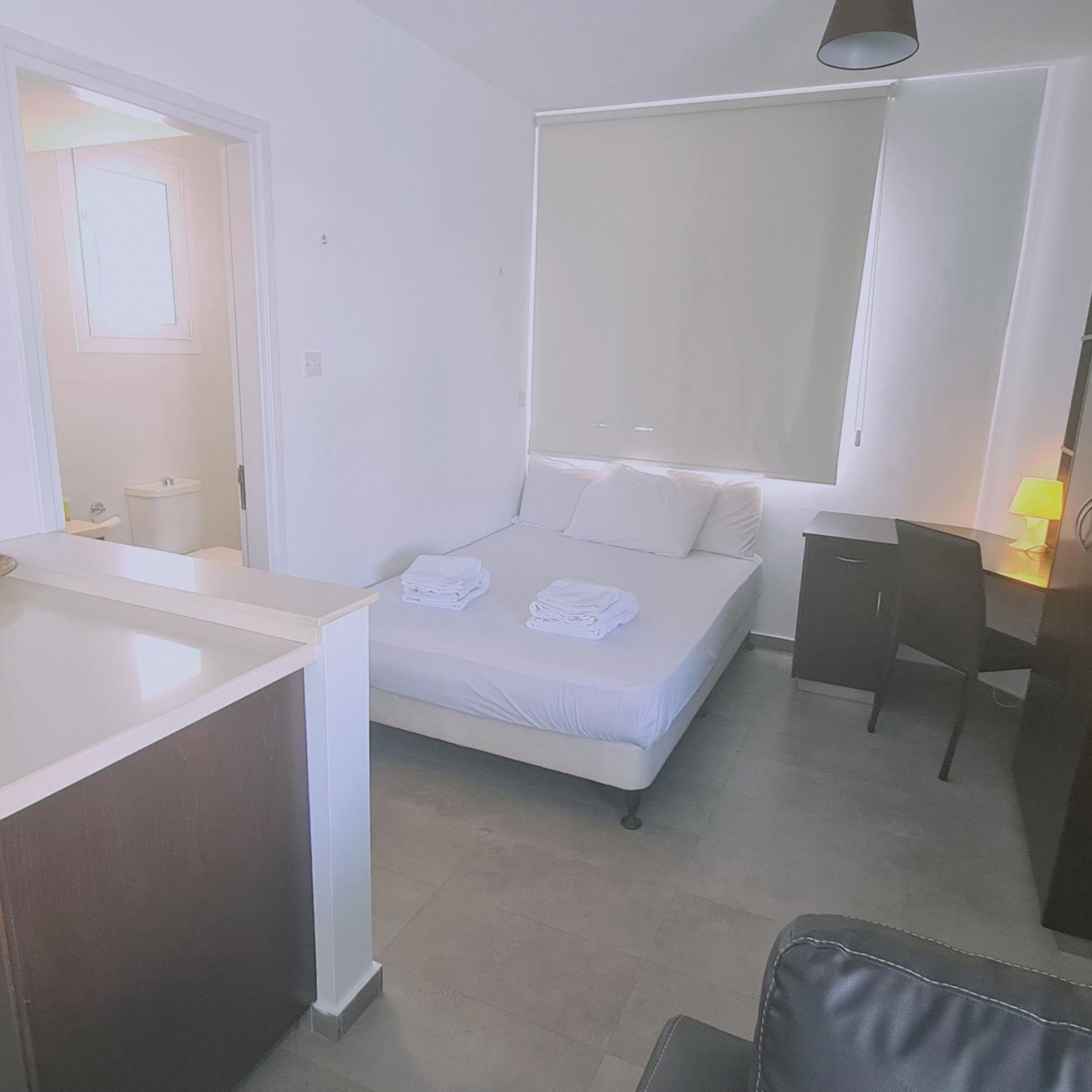 A small, modern bedroom with a single bed, a desk, and a chair. There is a window providing natural light and a view of the outside.