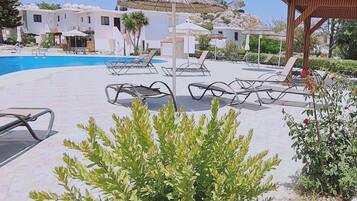 Un área junto a la piscina con sillones de descanso, un edificio pequeño con techo de paja y una pérgola de madera.