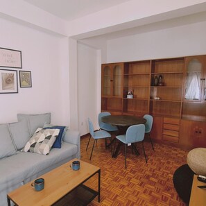 Un salon comprenant un canapé, une table basse et un coin repas avec une table ronde et des chaises. On y trouve également un meuble en bois avec des portes en verre et un miroir.