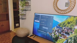 Ein Wohnzimmer mit Flachbildfernseher, der eine malerische Dorfsicht zeigt, einem Holzschrank, einem schwarzen Ottomanen und einem dekorativen Spiegel an der Wand.