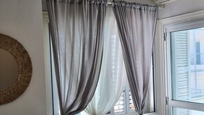 Ein Fenster mit grauen, zurückgebundenen Vorhängen, ein runder Spiegel an der Wand und eine weiße Klimaanlage darüber.