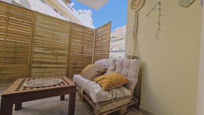 A cozy outdoor seating area with a wooden table and a cushioned bench, a woven mat on the table, and a wall-mounted light fixture. - 103 LCA - Stay Green Apartment (Larnaca)
