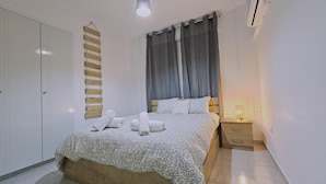 A bedroom with a bed, a bedside table with a lamp, a hanging wooden rack, and a wardrobe. - 103 LCA - Stay Green Apartment (Larnaca)