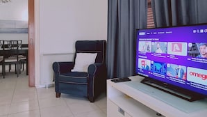 A living room with a flat-screen TV on a stand, a dark blue armchair, and a dining area with a table and chairs. - 75 NIC - Stay Mythos Down Town House (Nicosia)