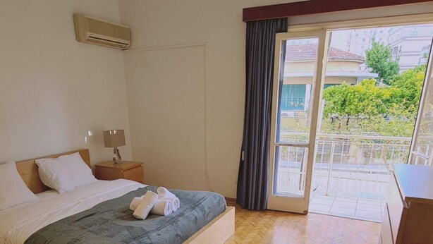 A hotel room with a wooden bed, a nightstand, a lamp, and a view of the outdoors through a sliding glass door. - 75 NIC - Stay Mythos Down Town House (Nicosia)