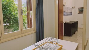 A room with a table, a window with curtains, a door leading to another room, and a view of greenery outside. - 75 NIC - Stay Mythos Down Town House (Nicosia)