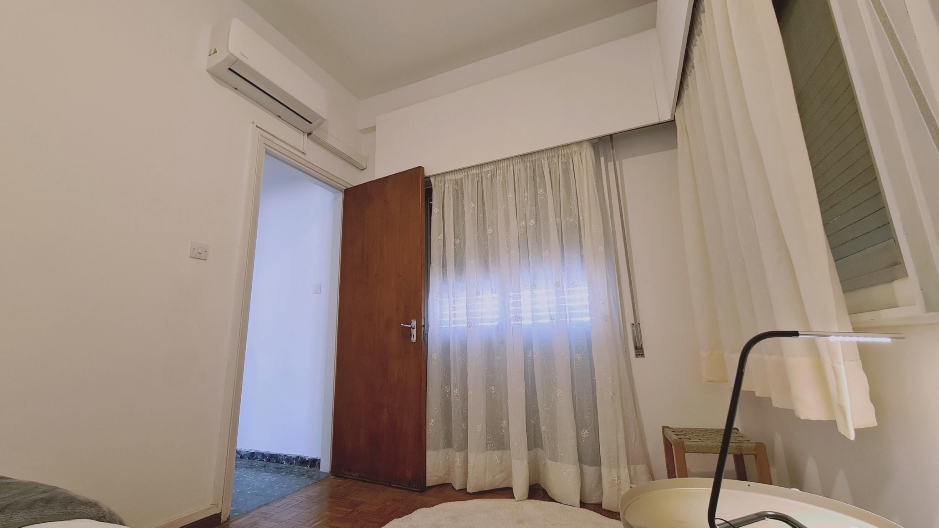 A room with a wooden door, a white curtain, a small table, and an air conditioning unit on the wall.