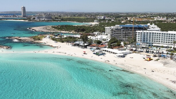 A coastal resort with a beachfront, a tall tower in the background, and a large hotel building. - 116 AN - Stay Nissi Blossom Apartment (Ayia Napa)