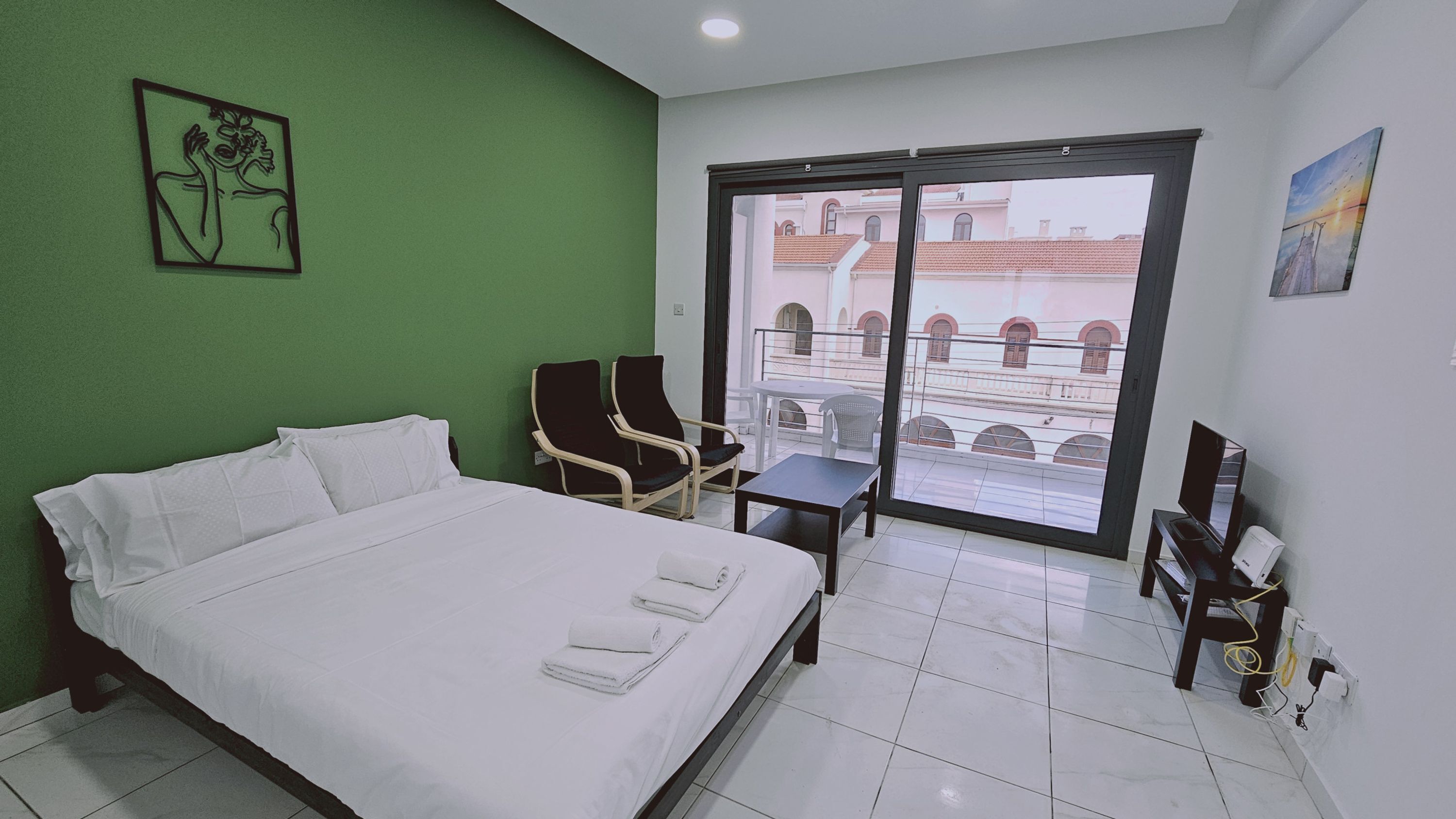 A hotel room with a bed, two chairs, a small table, and a large window with a view of buildings.