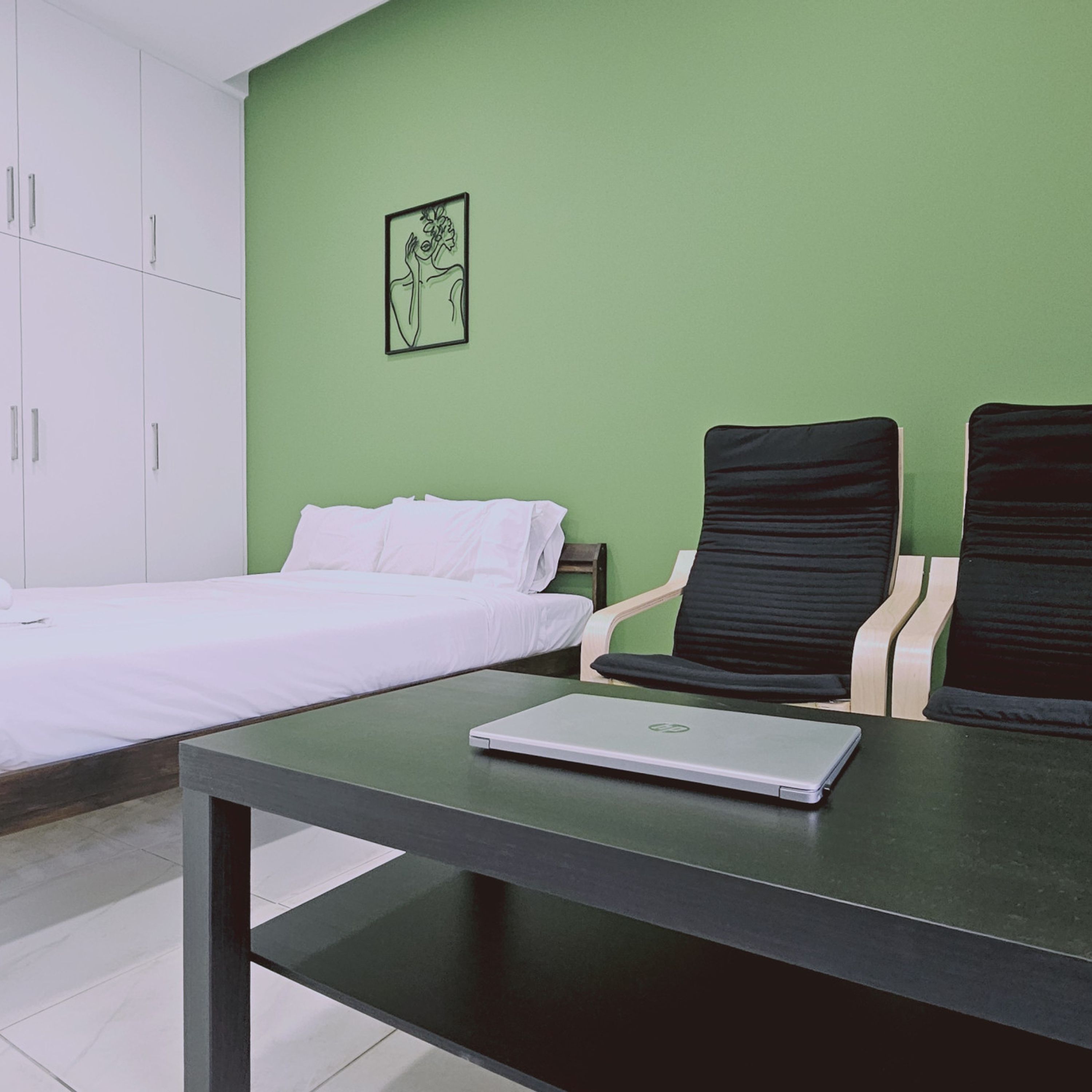 A hotel room with a bed, two black chairs, a table with a laptop, and a green wall.