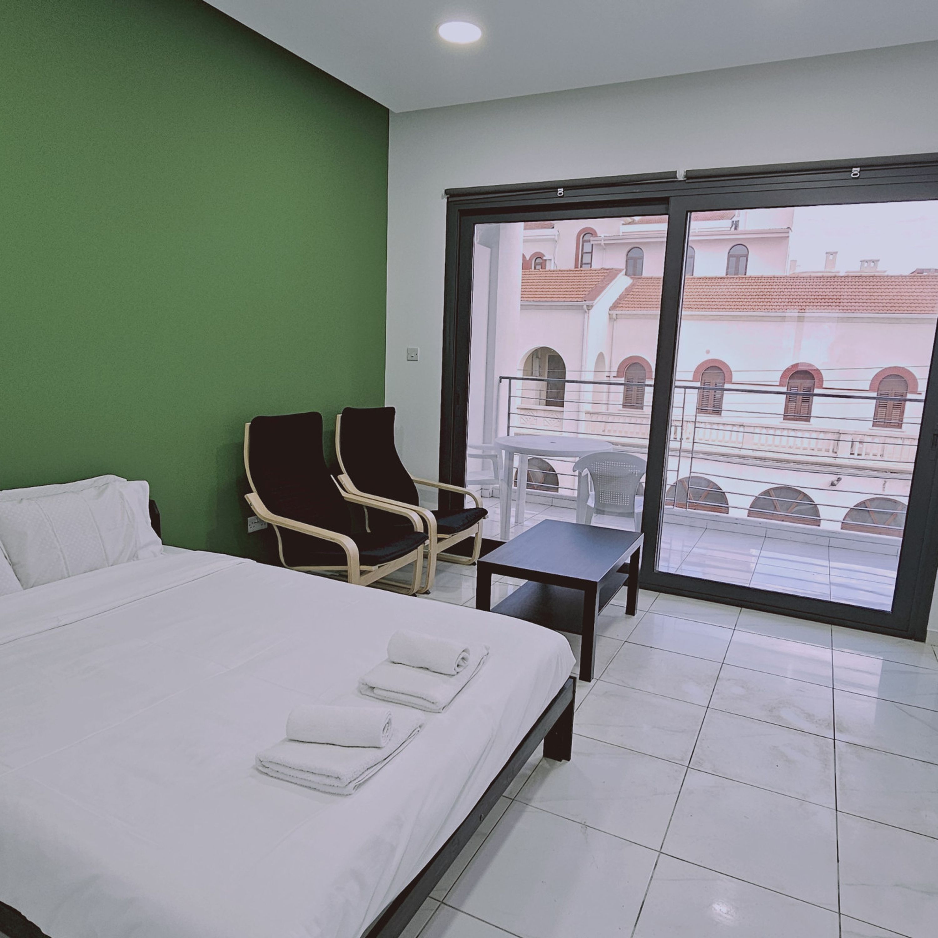 Quarto de hotel com cama, duas cadeiras, uma mesinha e varanda com vista para edifícios.
