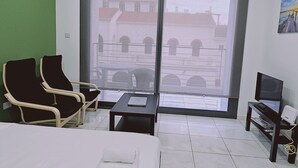 A hotel room with a bed, two chairs, a small table, a TV, and a large window with a view of buildings. - 109 LIM 1 - Stay Olive Studio (Limassol)
