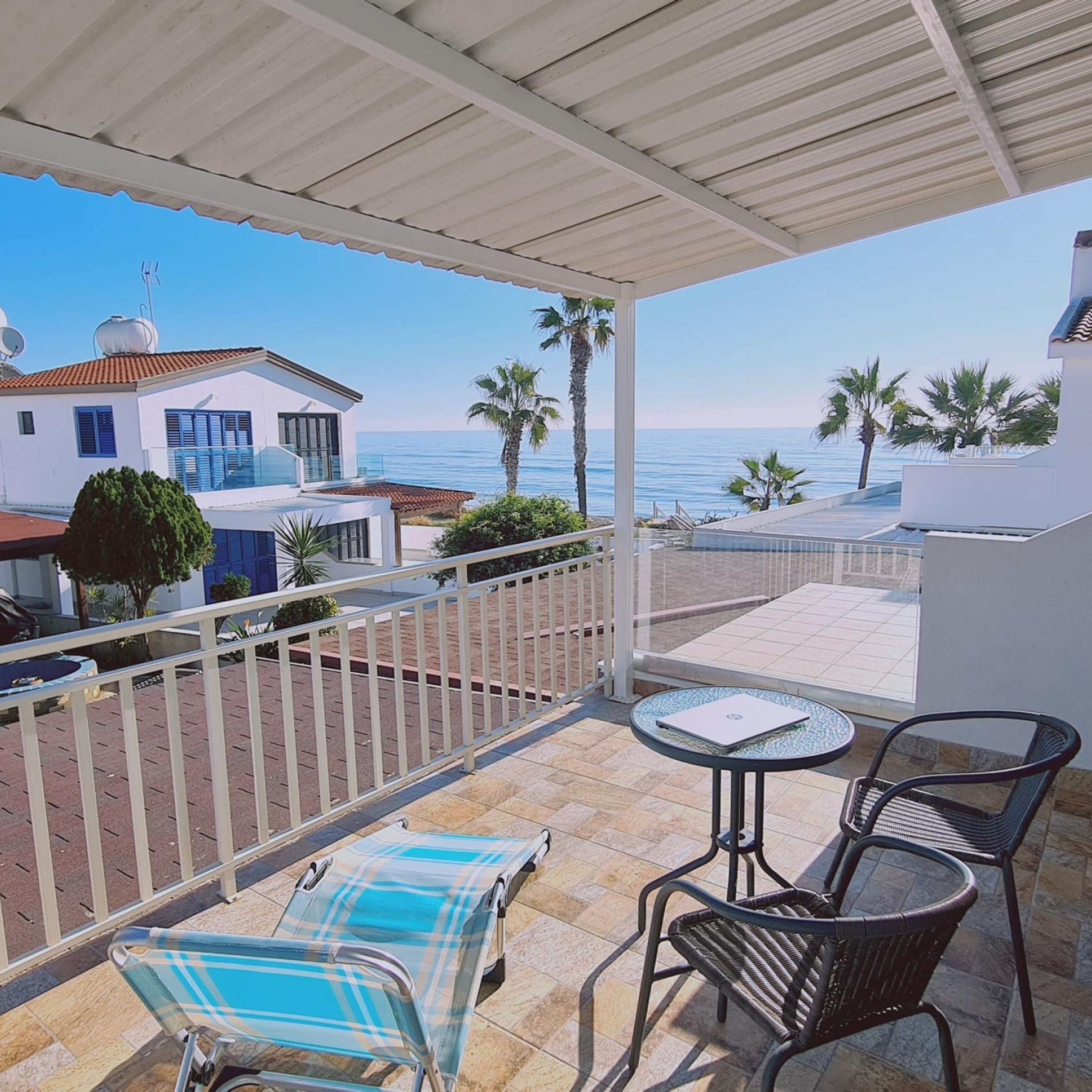 A covered outdoor area with a view of the sea, a lounge chair, a table, and a railing.
