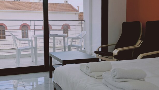 A hotel room with a bed, two chairs, a small table, and a view of buildings through large windows. - 109 LIM 3 - Stay Terra Cota Studio (Limassol)
