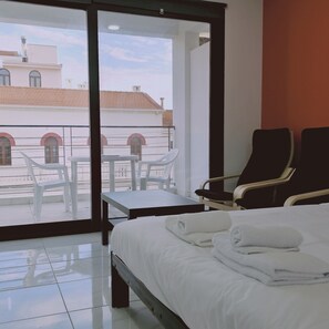 Habitación de hotel con cama, dos sillas, una mesita y vistas a edificios a través de amplias ventanas.