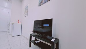 A modern living room with a flat-screen TV on a black stand, a white refrigerator, and a small table with a remote control. - 109 LIM 3 - Stay Terra Cota Studio (Limassol)